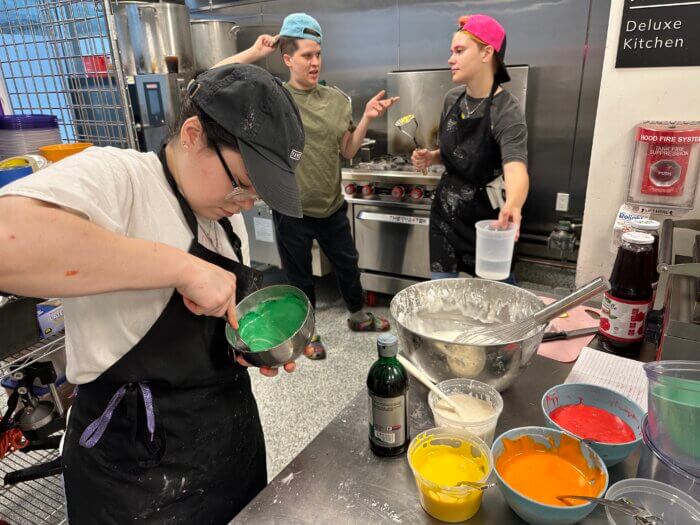 The Bite team in the kitchen making rainbow icing to decorate their signature Gaydonut. (Pictured L to R) Maeve mixes rainbow colors as B and Maya discuss other flavors.