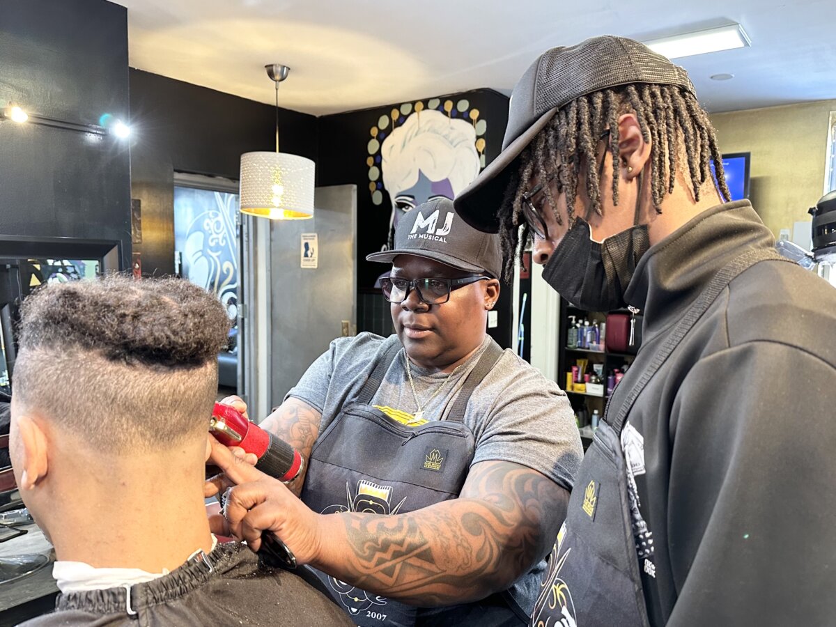 Khane Kutzwell (left) mentors apprentice barber Da’Shawn Scott (right) while touching up the sides on a Fordham Law School student.