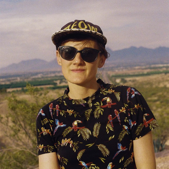 Alessandra Lacorazza wears sunglasses while smiling for a headshot against a backdrop of natural landscape.