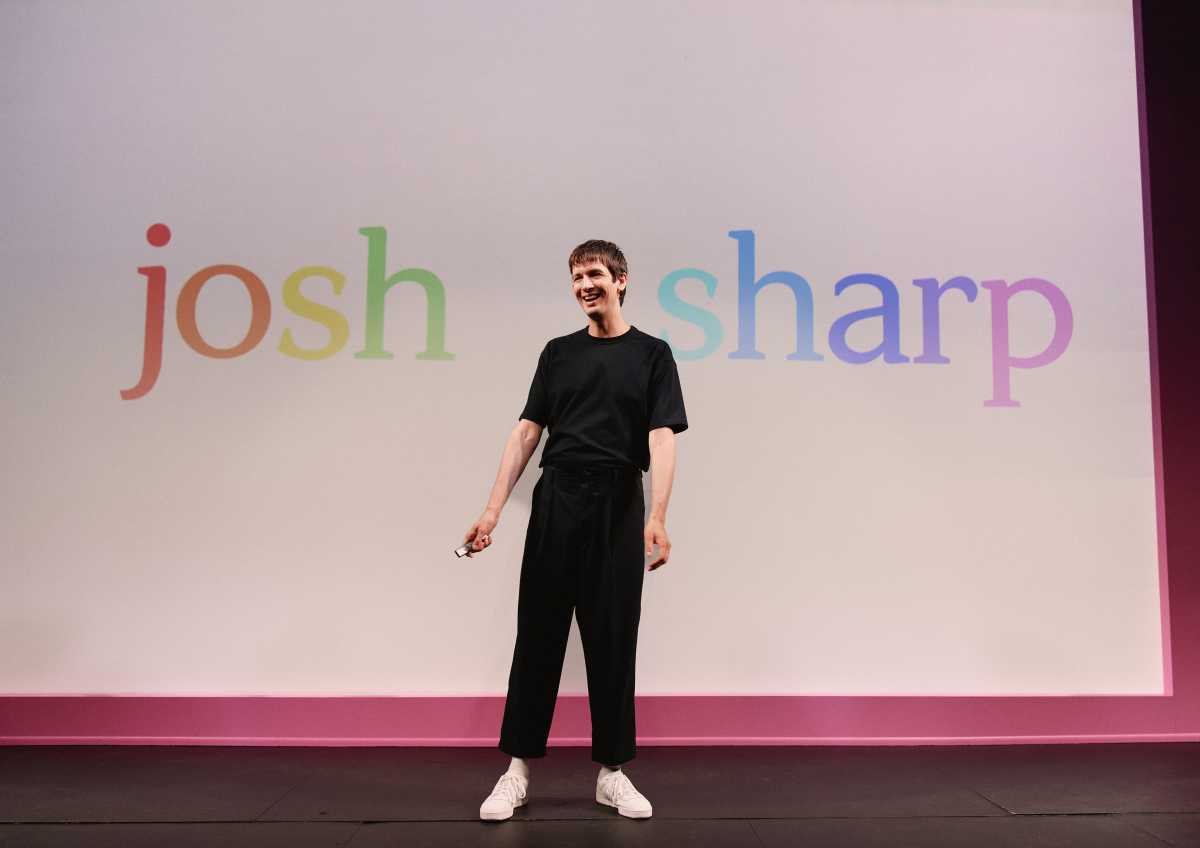 Josh Sharp stands on stage with his name printed on screen behind him in rainbow colors.