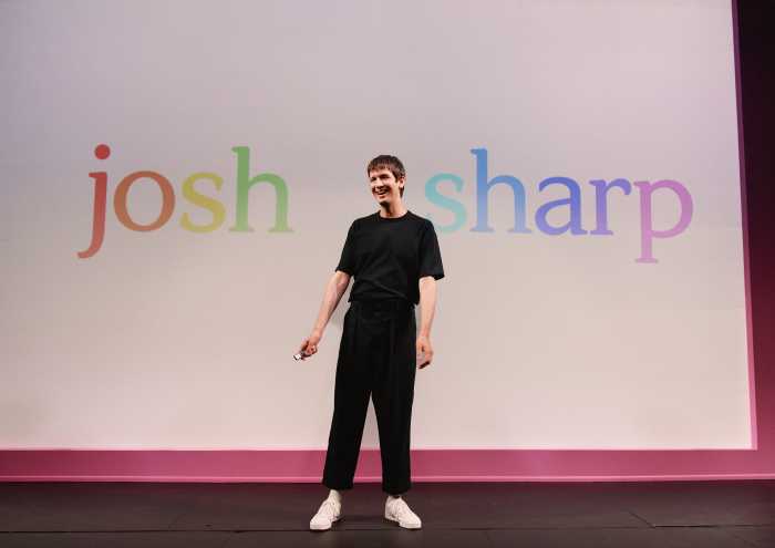 Josh Sharp stands on stage with his name printed on screen behind him in rainbow colors.