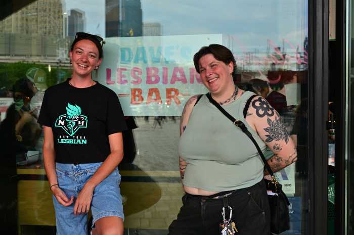 Two people stand in front of a window with a sign advertising Dave's Lesbian Bar.