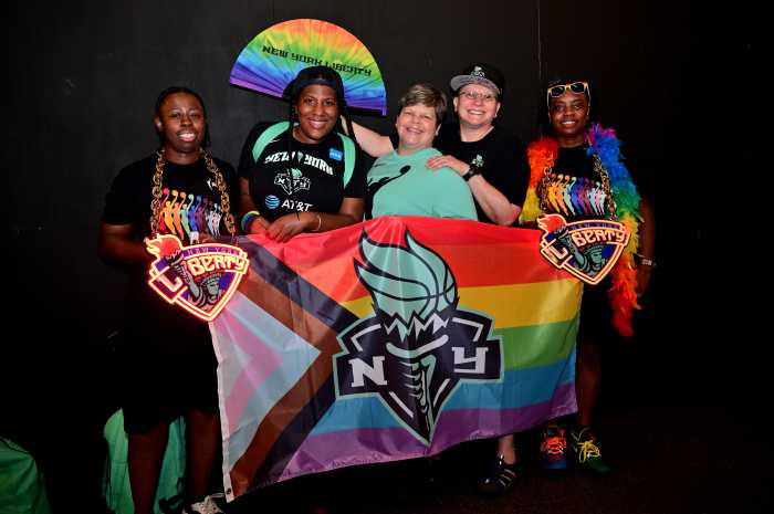 Five Liberty fans stand together in front of a Rainbow Flag with a New York Liberty logo.