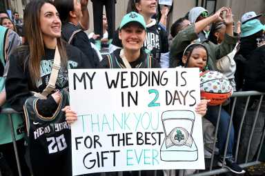 An individual holds a sign that reads: "My wedding is in 2 days. Thank you for the best gift ever." The sign features a New York Liberty championship ring. The subject is smiling while surrounded by the crowd.